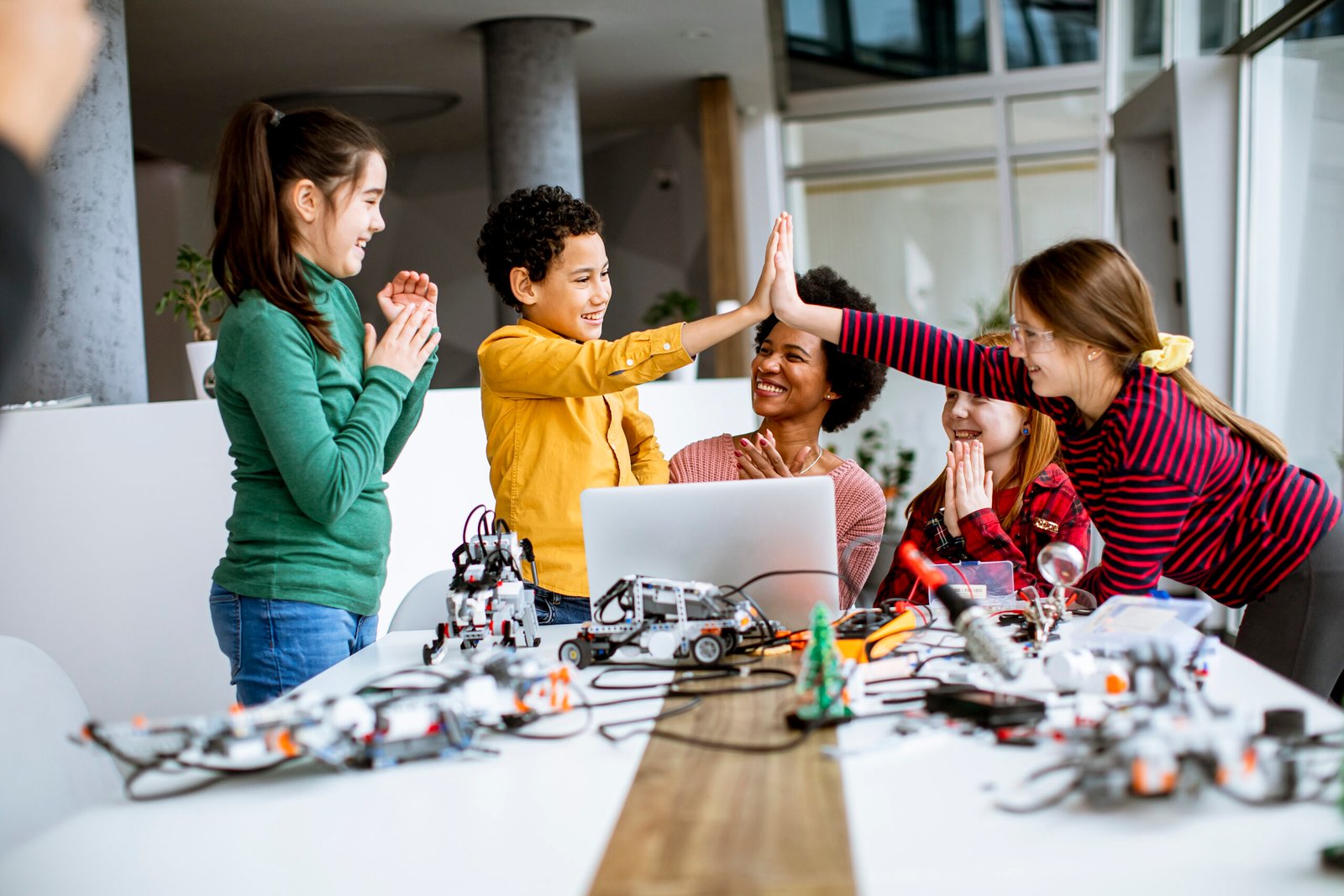 Group of happy kids with their African American female science teacher with laptop programming electric toys and robots at robotics classroom